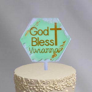 以下が含まれることがあります： 緑色の背景に金色の十字架と「God Bless Vivianna」という金色の文字が書かれた六角形のケーキトッパーが飾られた、バタークリームでデコレーションされた2段の白いケーキです。