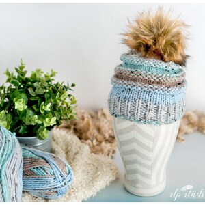 K&ouml;nnte beinhalten: Eine gestrickte M&uuml;tze in Blau- und Braunt&ouml;nen mit einem flauschigen braunen Bommel oben. Die M&uuml;tze wird auf einem wei&szlig;en und grauen Mannequin-Kopf pr&auml;sentiert.