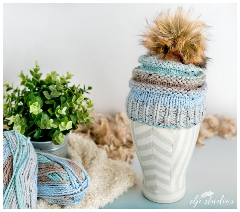 K&ouml;nnte beinhalten: Eine gestrickte M&uuml;tze in Blau- und Braunt&ouml;nen mit einem flauschigen braunen Bommel oben. Die M&uuml;tze wird auf einem wei&szlig;en und grauen Mannequin-Kopf pr&auml;sentiert.
