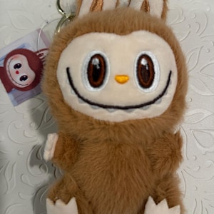 May include: A plush brown bunny keychain with a smiling face, large brown eyes, and small white paws. The keychain has a gold-colored metal ring and a small tag with a cartoon character.