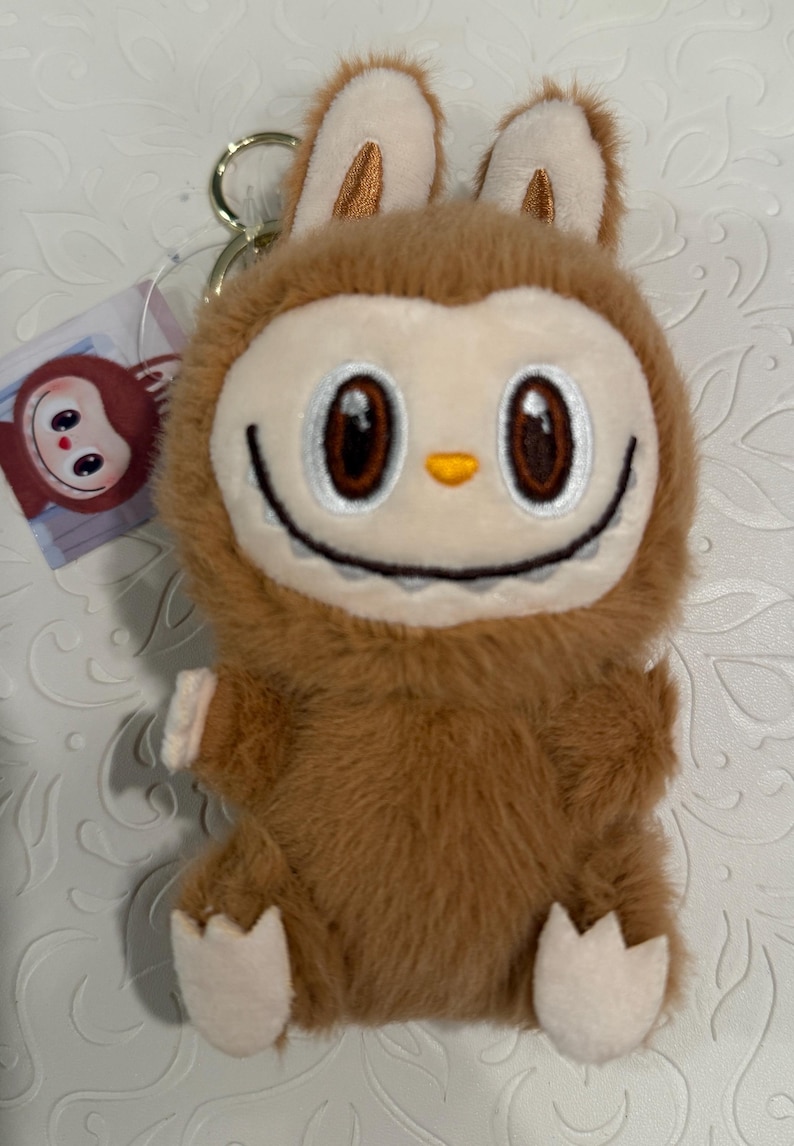 May include: A plush brown bunny keychain with a smiling face, large brown eyes, and small white paws. The keychain has a gold-colored metal ring and a small tag with a cartoon character.