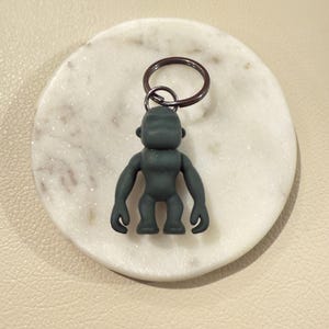 May include: A dark gray gorilla keychain. The gorilla figure has a stylized design with a round head, short arms, and legs. The keychain ring is dark brown. The gorilla is resting on a white marble coaster.