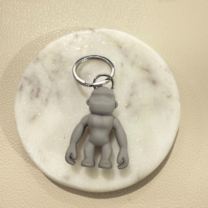 May include: A gray gorilla keychain with a silver keyring. The gorilla figure is standing upright with its arms at its sides. The keychain is resting on a white and gray marble coaster.