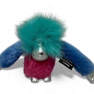May include: A plush toy in the shape of a creature with a teal furry head, blue arms, and a magenta body. The creature has silver-colored hands, feet, and face. A black tag reads "QUENTIN" and "www.kipling.com".