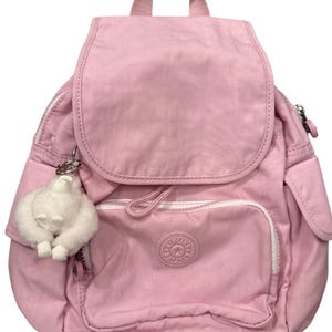 May include: A light pink backpack with a flap closure and a small front pocket. The backpack has a top handle and adjustable straps. A white fluffy monkey keychain hangs from the zipper. The brand logo is visible on the front pocket.