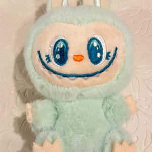 May include: A plush toy in the shape of a cartoon creature with a bunny-like head. The toy is primarily light blue with a cream-colored face, ears, hands, and feet. It has large blue eyes, a small orange nose, and a smiling mouth.