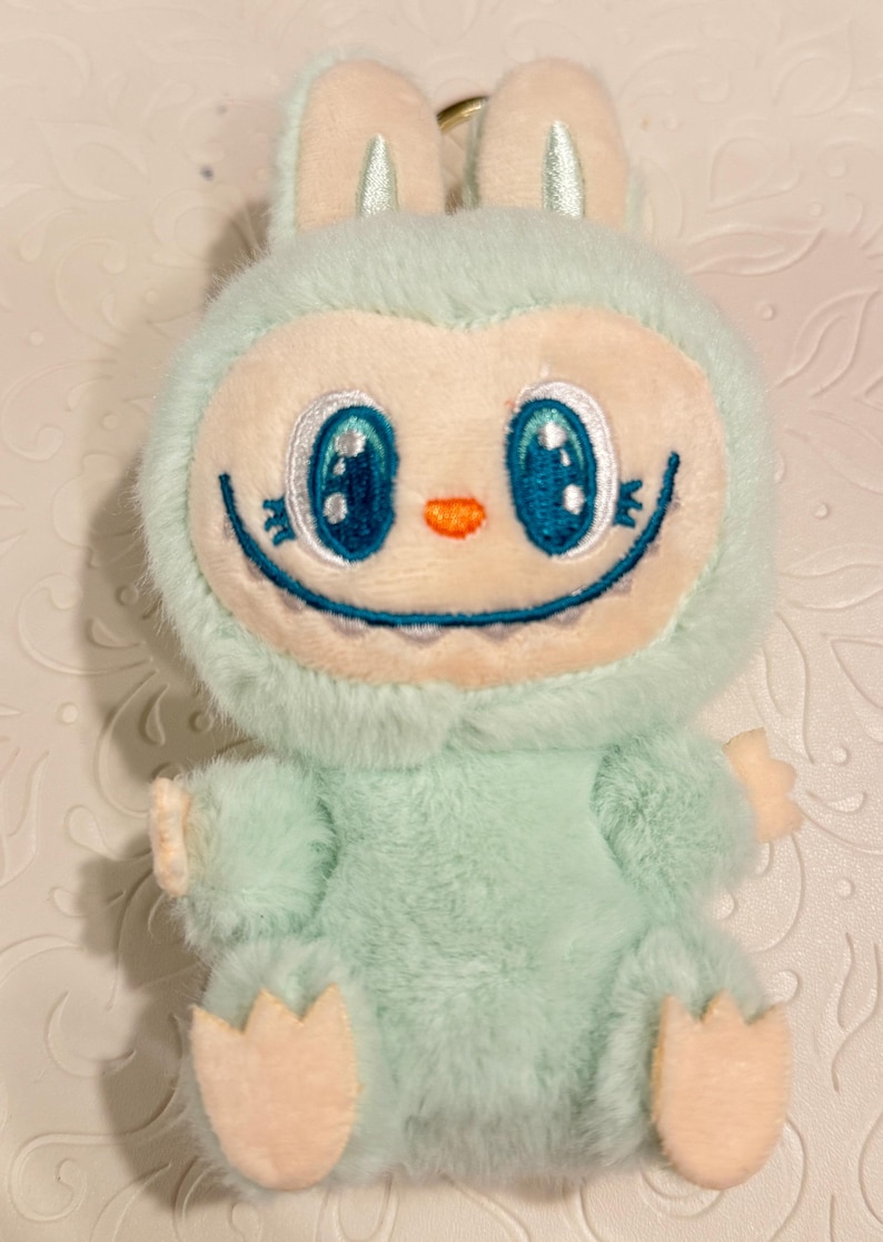 May include: A plush toy in the shape of a cartoon creature with a bunny-like head. The toy is primarily light blue with a cream-colored face, ears, hands, and feet. It has large blue eyes, a small orange nose, and a smiling mouth.