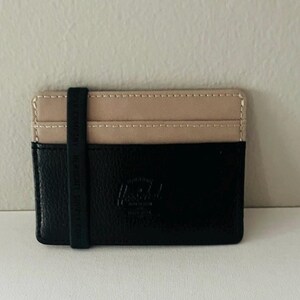 May include: A black and tan cardholder with a black elastic band. The cardholder is made of a textured black material on the bottom half and tan canvas on the top half. The elastic band has white text. The brand logo is embossed on the black section.