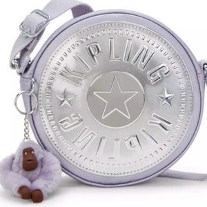 May include: A silver and purple round crossbody bag with a silver Kipling logo and a star. The bag has a furry purple monkey keychain.