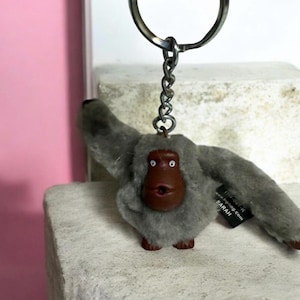 May include: A gray plush monkey keychain with a brown face and outstretched arms. The keychain has a metal ring and chain. A small black tag with white text is attached to the keychain.