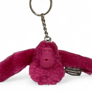 May include: A magenta plush monkey keychain with outstretched arms. The monkey has a textured body and a smooth face. A small tag with text is attached to the monkey.