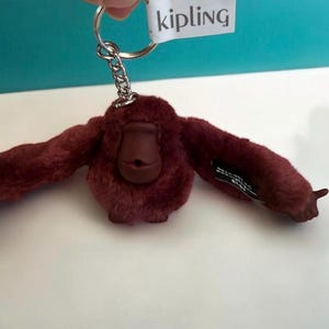 May include: A maroon-colored plush orangutan keychain with a silver keyring. The keychain has a brown face and outstretched arms. The tag reads "kipling".