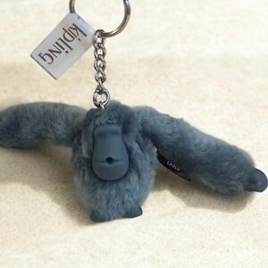 May include: A blue plush monkey keychain with a silver keyring and chain. The monkey has outstretched arms and a dark blue face. A white tag with the word "Kipling" is attached to the keyring.