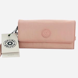 Puede incluir: Una cartera rosa Kipling con cierre de cremallera y una correa desmontable. La cartera tiene un logotipo de Kipling con una estrella en la parte delantera.