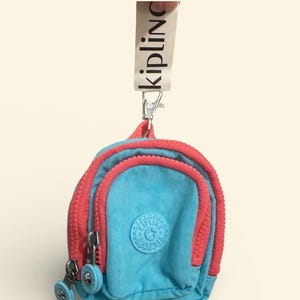 May include: A small, turquoise blue backpack-shaped pouch with a coral pink zipper and trim. The pouch has a circular logo and the word "Kipling" printed on a white strap attached to a silver clasp.