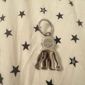 May include: A silver-colored keychain featuring a stylized figure with multiple limbs and a circular emblem. The keychain is attached to a metal ring. The background is a white fabric with a pattern of black stars.