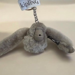 May include: A gray plush monkey keychain with a silver chain and a tag that reads "Kipling". The monkey has outstretched arms and a small, round face. The keychain is designed to be attached to keys or a bag.