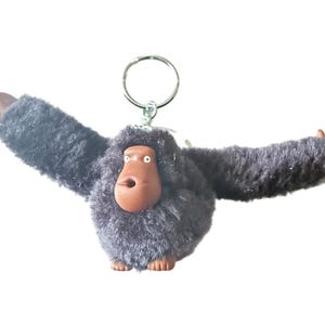 May include: A plush gorilla keychain with outstretched arms and a thumbs-up gesture. The gorilla has a brown face and hands, with a dark gray, shaggy body. A silver keyring is attached to the top of the gorilla's head.