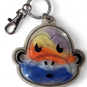May include: A silver-toned keychain featuring a monkey face design with a colorful, textured interior. The monkey's face is outlined in silver, with black eyes and a nose. The interior is filled with yellow, orange, purple, and blue fibers. A silver keyring and clasp are attached.
