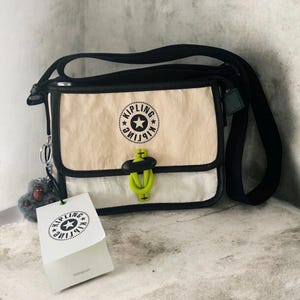 May include: A black and beige crossbody bag with a black strap. The bag features a flap closure with a neon green accent and a logo. A small gray keychain and a tag with a logo are attached.