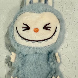 May include: A plush, light blue creature-shaped keychain with a smiling face, small arms, and feet. The face is white with black eyes and a toothy grin. The ears are light blue with silver accents.
