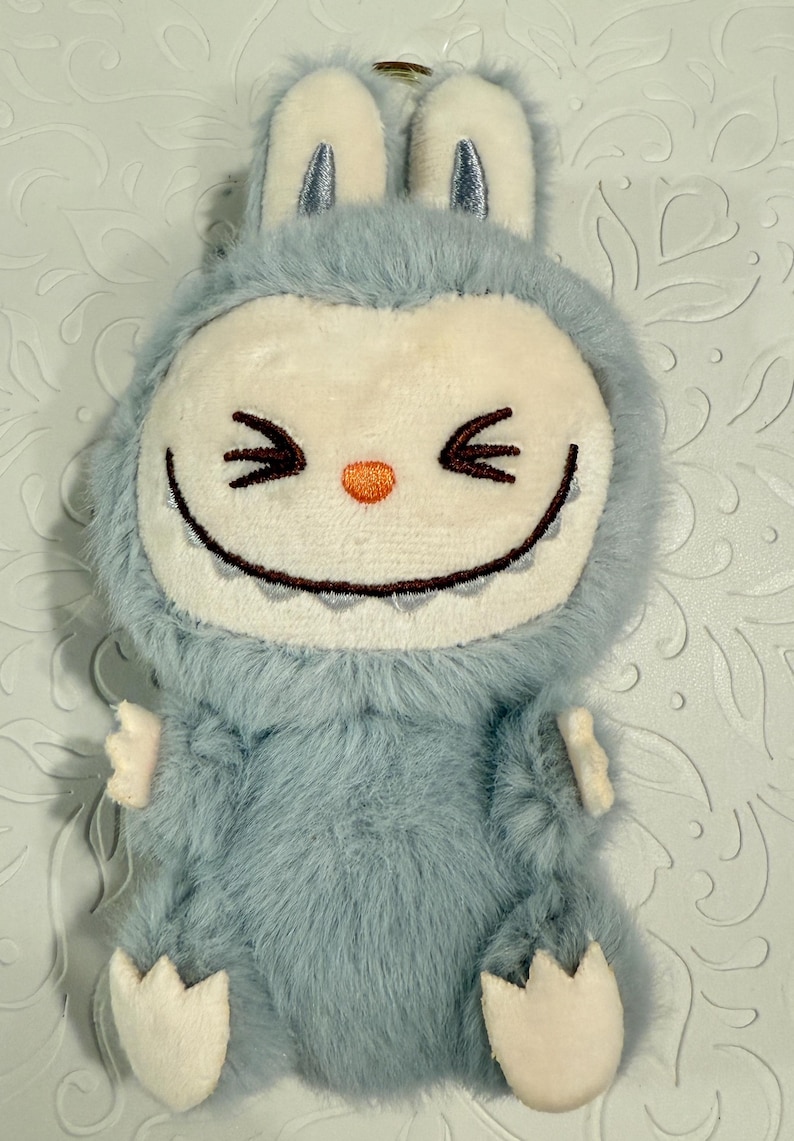 May include: A plush, light blue creature-shaped keychain with a smiling face, small arms, and feet. The face is white with black eyes and a toothy grin. The ears are light blue with silver accents.