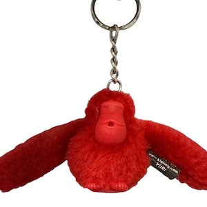 May include: A red plush monkey keychain with outstretched arms. The monkey has a red face and body, with a silver metal keyring attached. The tag reads "YURI" and "www.kipling.com".
