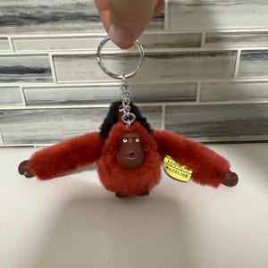 Peut inclure: Un porte-clés orang-outan en peluche rouge avec des cheveux noirs et des bras et un visage bruns. Le porte-clés a un anneau argenté et une petite étiquette qui dit "Kipling MADELINE".