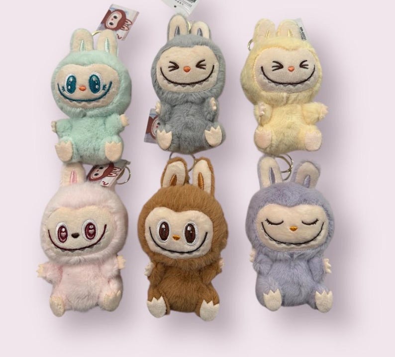 May include: Six plush bunny keychains in various colors: mint green, gray, yellow, pink, brown, and lavender. Each plush has a smiling face and a hood. The keychains are attached to small tags.