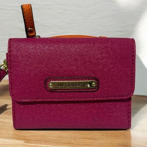 May include: A magenta Liz Claiborne wallet with a textured finish. The wallet has a gold-tone metal nameplate and zipper pull. It features a wrist strap and a contrasting orange handle. The wallet is sitting on a wooden surface.