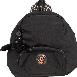 May include: Black nylon backpack with a small front pocket and a top handle with a geometric pattern. A small black monkey keychain hangs from the zipper. The backpack has a round logo with a star.