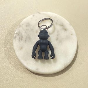 May include: A dark gray gorilla-shaped keychain. The keychain is made of a solid material and features a silver-colored key ring. The gorilla is in a squatting position with its arms at its sides. The keychain rests on a white marble coaster.