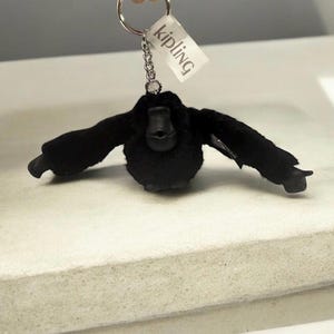 May include: A black plush gorilla keychain with outstretched arms. The keychain has a silver metal ring and chain, with a small white tag that reads "Kipling". The gorilla has a round body and a detailed face.