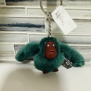 May include: A green plush monkey keychain with a brown face and a silver keyring. The keychain has a tag that says "Kipling". The monkey has a fluffy texture and is in a sitting position.