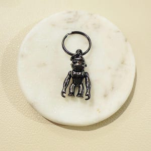 May include: A dark gray metal keychain featuring a small, articulated gorilla figure. The gorilla has a detailed design with visible arms, legs, and a head. The keychain is attached to a circular ring. The item is set on a white, circular surface.