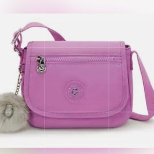 May include: A lavender crossbody bag with a flap closure, a zippered pocket, and a long adjustable strap. The bag features a silver charm and a fluffy, light gray pom-pom. The bag is made of a smooth, durable material.