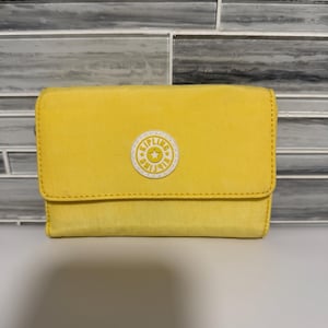 Può includere: Un portafoglio giallo brillante con chiusura a patta. Il portafoglio ha un logo circolare bianco con la scritta "Kipling" e una stella al centro. Il portafoglio è realizzato in tessuto strutturato. Misura circa 12 cm x 9 cm.