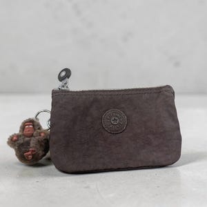 May include: A brown nylon coin purse with a zipper closure and a small, round logo. A brown monkey keychain is attached to the side. The purse is approximately 5 inches wide and 3 inches tall.