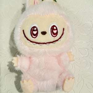 May include: A plush pink keychain featuring a cartoon character with a smiling face, red heart-shaped eyes, and a small nose. The character has a furry body, small arms and feet, and bunny-like ears. A gold-colored ring is attached to the top.
