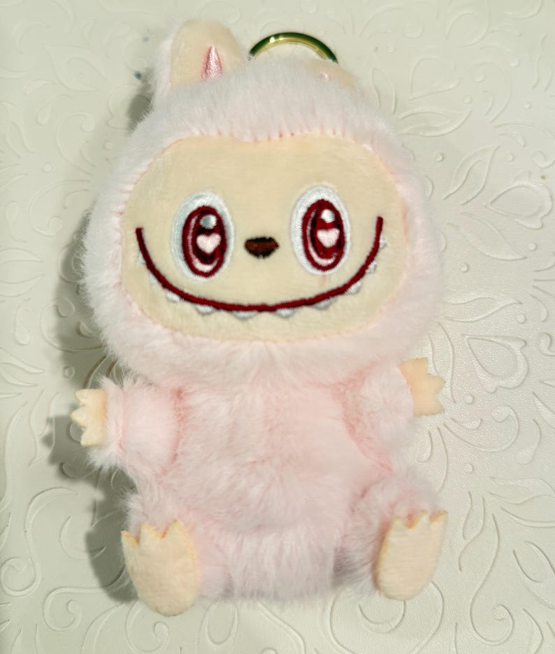 May include: A plush pink keychain featuring a cartoon character with a smiling face, red heart-shaped eyes, and a small nose. The character has a furry body, small arms and feet, and bunny-like ears. A gold-colored ring is attached to the top.
