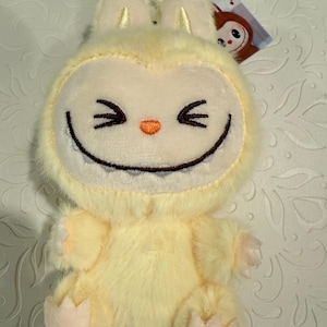 May include: A plush, light yellow creature with a smiling face, pointed ears, and small arms and feet. The creature has black stitched eyes and a wide, toothy grin. A small tag with a cartoon character is attached.