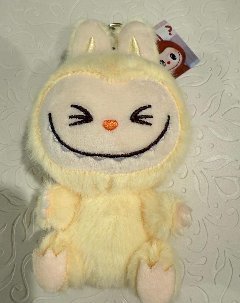 May include: A plush, light yellow creature with a smiling face, pointed ears, and small arms and feet. The creature has black stitched eyes and a wide, toothy grin. A small tag with a cartoon character is attached.
