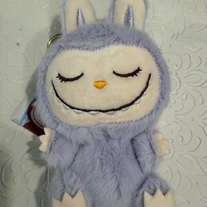 May include: A plush, lavender-colored bunny keychain with a smiling face and closed eyes. The bunny has a white face, ears, and paws, with a small orange nose. The keychain is made of soft, fuzzy material.