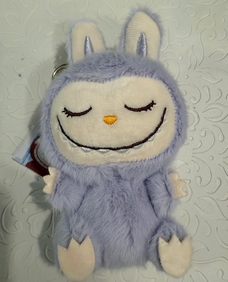 May include: A plush, lavender-colored bunny keychain with a smiling face and closed eyes. The bunny has a white face, ears, and paws, with a small orange nose. The keychain is made of soft, fuzzy material.