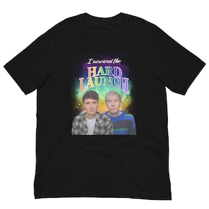 May include: Black t-shirt featuring two men and the text "I survived the HARD LAUNCH" in a colorful, stylized font. One man wears a plaid shirt, the other a blue and green striped sweater.