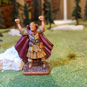 May include: A painted miniature figurine of a man wearing a red and brown robe, with his arms raised in the air. He is wearing a blue amulet around his neck and a brown belt with a pouch attached. The figurine is standing on a brown base.