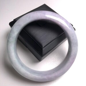 Puede incluir: Un brazalete de jade pulido con un degradado de tonos lavanda y verde pálido. El brazalete liso y circular se exhibe sobre una caja negra, mostrando su acabado brillante y sus variaciones de color naturales. Una joya.