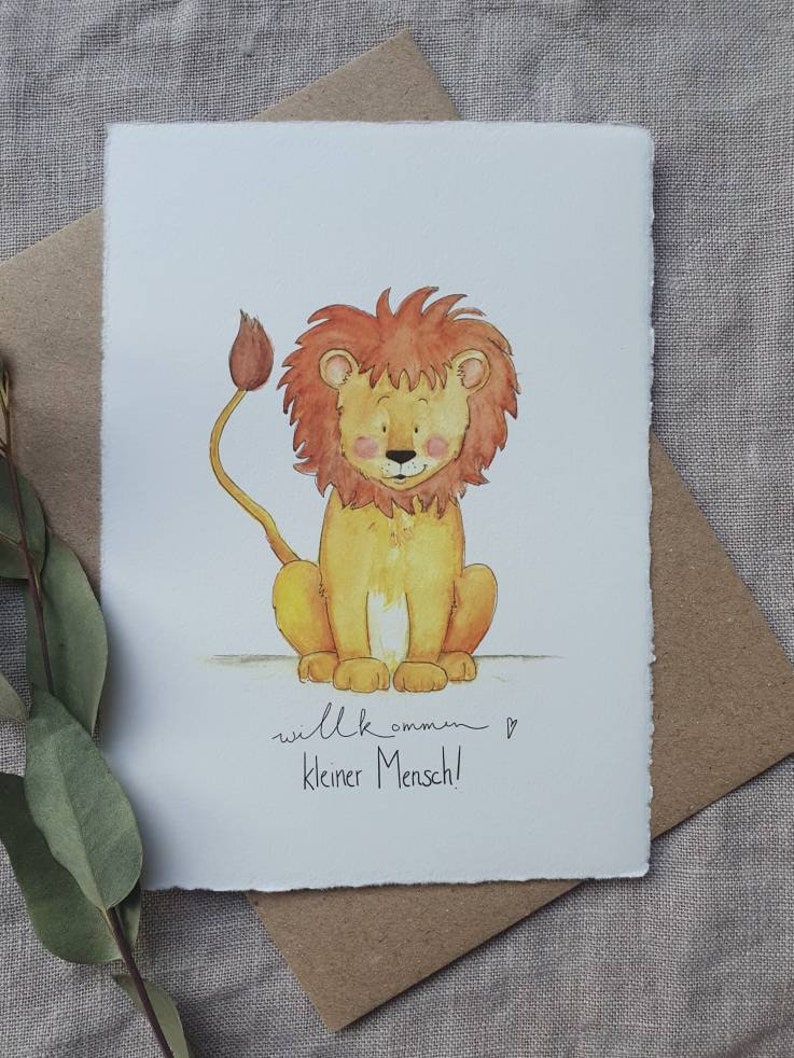 May include: A watercolor illustration of a cute lion with a red mane and a yellow body. The lion is sitting down and looking at the viewer. The text "willkommen kleiner Mensch!" is written below the lion.