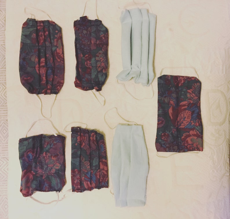 May include: Six face masks, three with a floral pattern in shades of red, black, and white, and three plain white masks. The masks are hanging on a light beige surface.