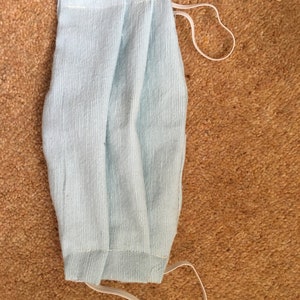 May include: A light blue fabric face mask with white elastic ear loops. The mask is folded in half and has a pleated design.